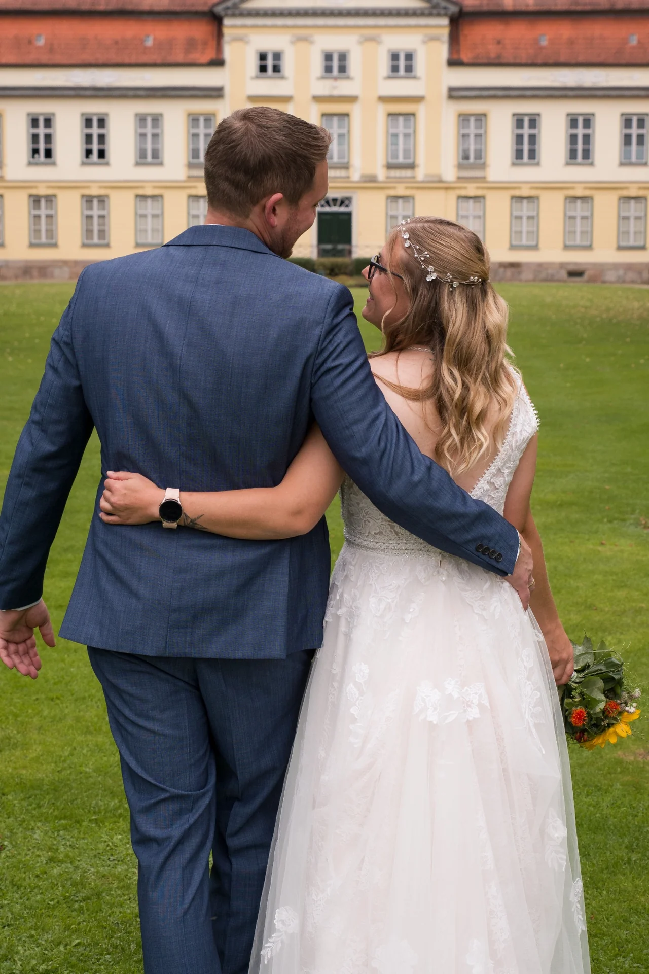 Das Paar steht Arm in Arm vor einem Gutshaus, von hinten fotografiert, und schaut sich an.