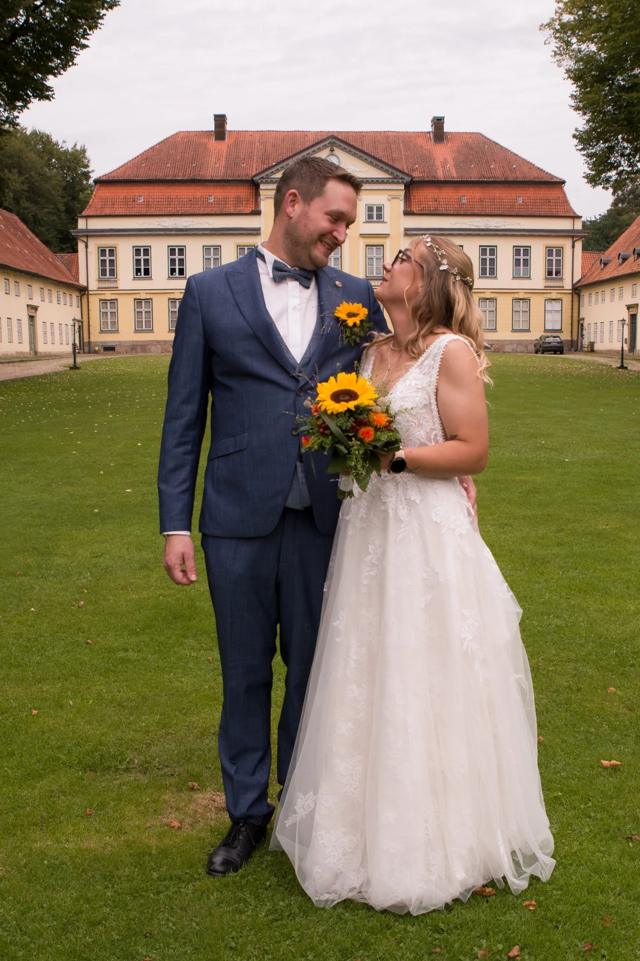 Braut und Bräutigam auf der Wiese vor einem gelben Gutshaus mit rotem Dach.